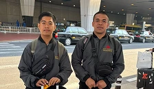Dua pria mengenakan jaket kerja dan tas selempang duduk berdampingan di area publik.